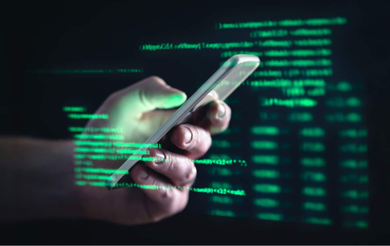 Imagem mostra uma mão segurando um smartphone e, ao redor dela, uma série de códigos e dados em fonte verde, simbolizando o acesso a informações privadas e golpes cibernéticos