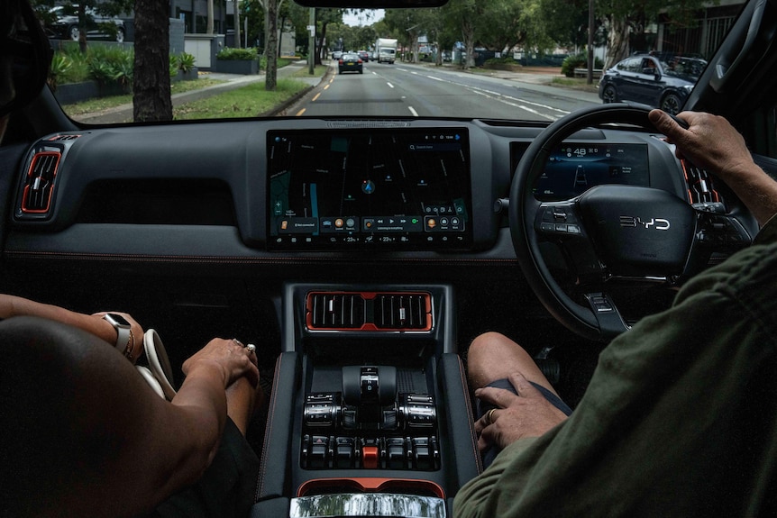 A driver and passenger sit in a modern BYD car driving on the road. The photo is taken from the back seat.