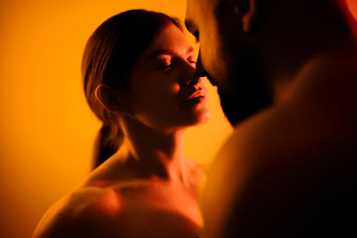 Casal em momento íntimo próximos e quase se beijando. A imagem é colorida e eles estão em um fundo alaranjado - Metrópoles