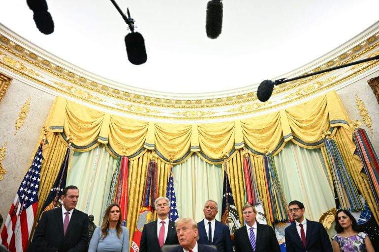 Donald Trump stands among a group of officials in dark suits in the Oval Office, in front of tall gold curtains.