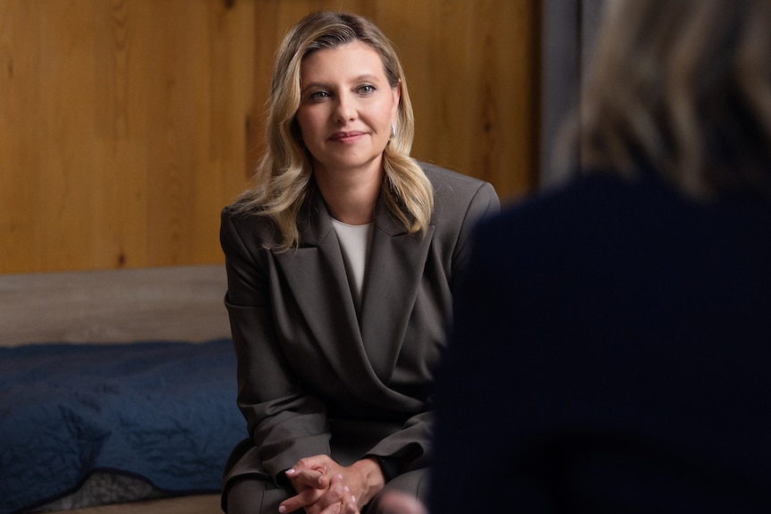 A women with blonde hair and wearing an olive-green suit is being interviewed.